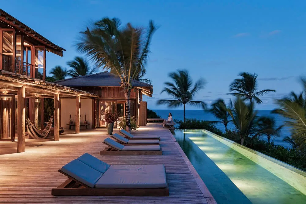 Illuminated resort pool deck at dusk with timber loungers, palms and guest buildings