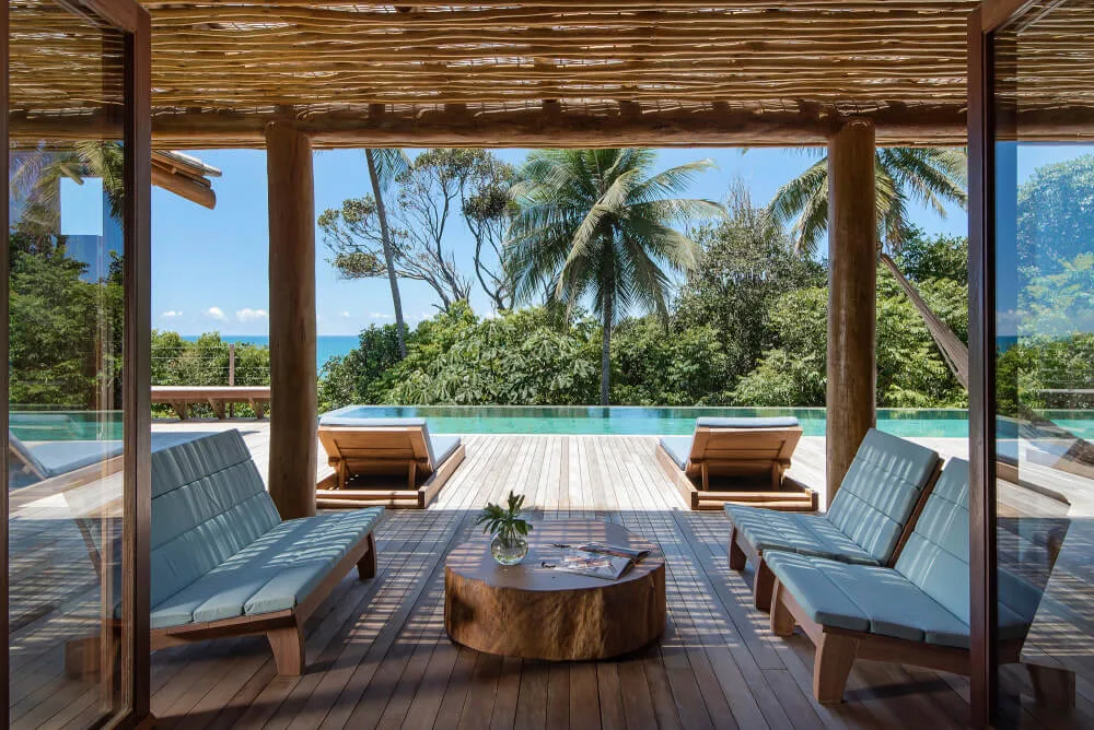 Covered lounge with timber chairs opening directly to a pool terrace and tropical vegetation