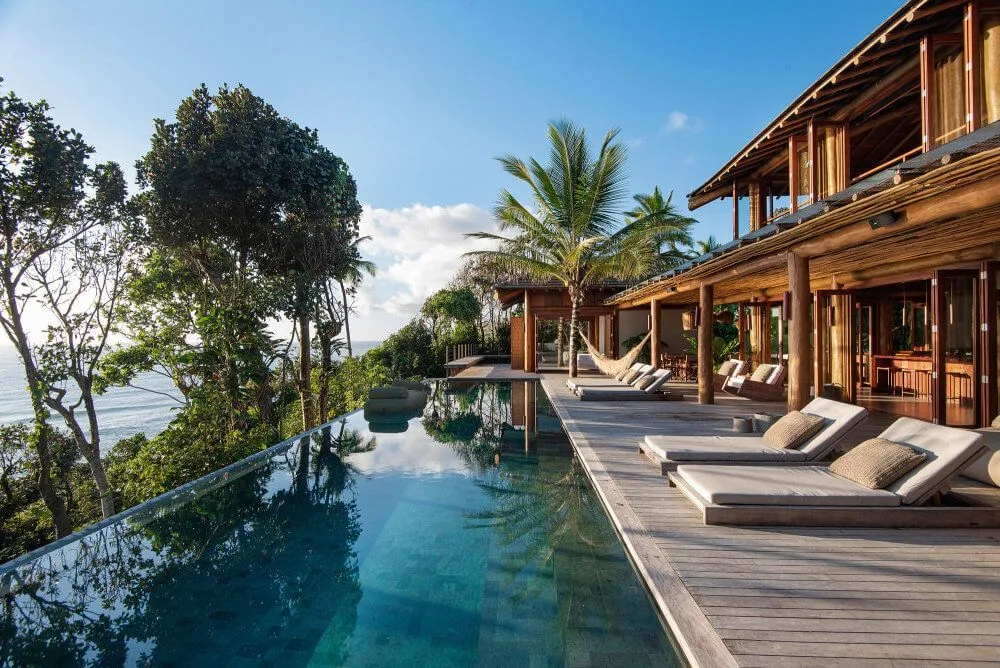Long swimming pool beside a timber deck with a row of cushioned loungers and ocean-facing resort rooms